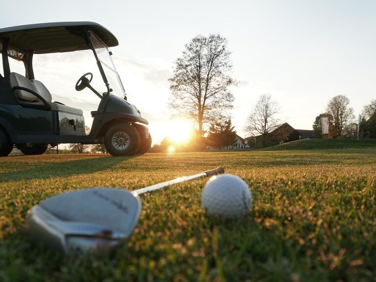 Ein Golfschläger, Golfcart und Golfball werden bei Sonnenaufgang in Szene gesetzt.