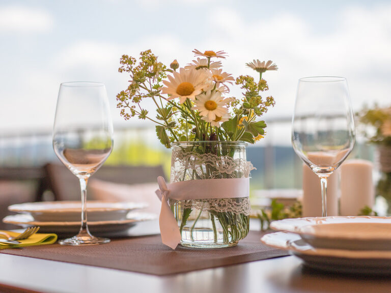 Auf der Sonnenterrasse steht ein liebevoll gedeckter Tisch mit feinem Weinservice aus Glas und einer Blumenvase als dekorativem Mittelpunkt.